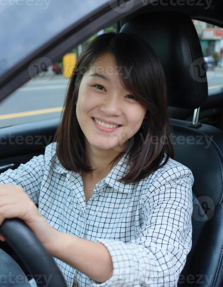 A smiling Asian woman with shoulder-length brown hair drives a car, wearing a white shirt photo