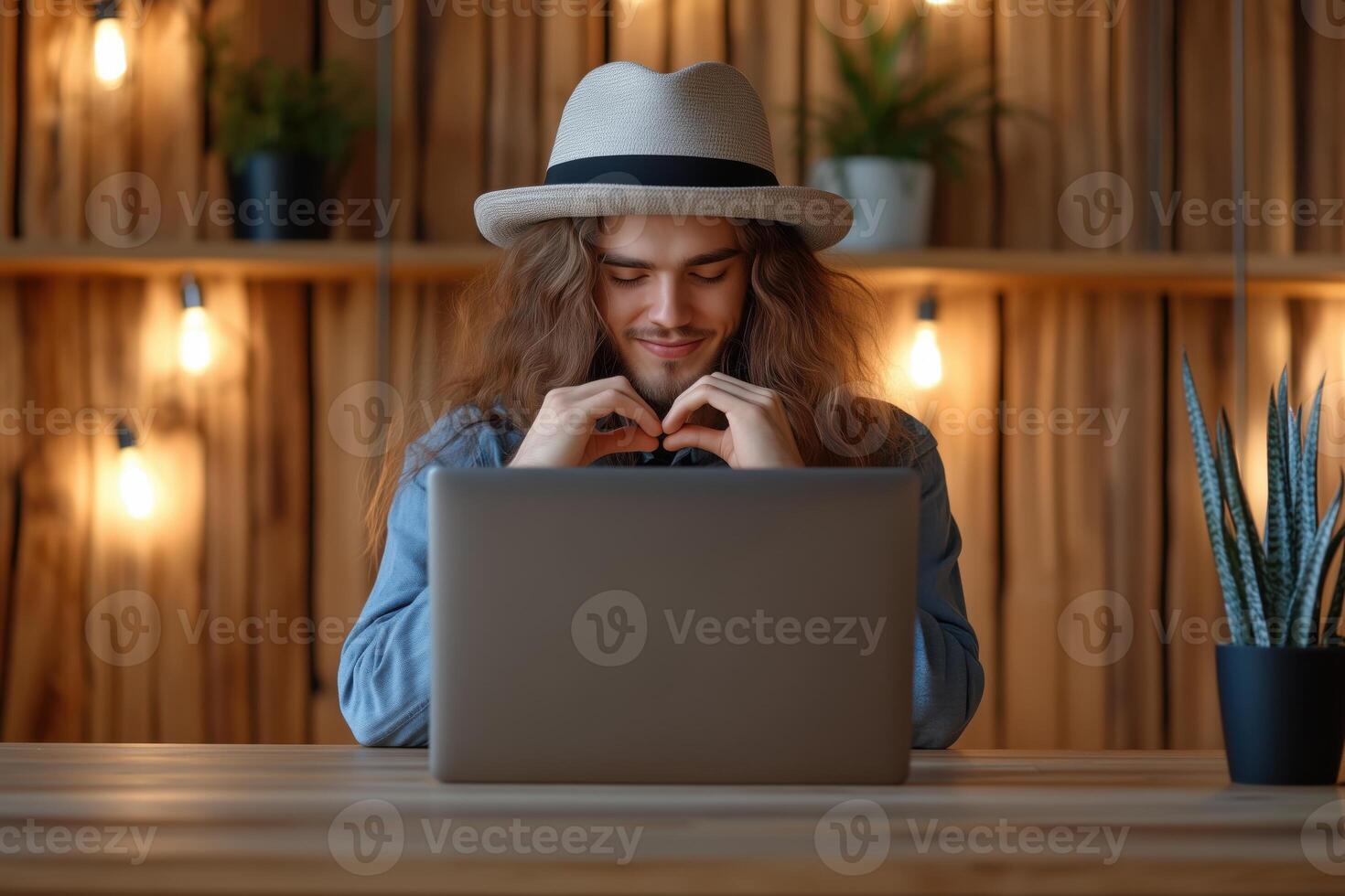 Man with long hair and hat forms a heart with hands during a call at a cozy wooden table photo
