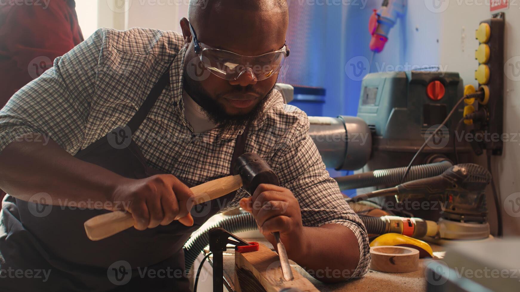 African american sculptor shaping raw timber using chisel and hammer in carpentry shop, creating wood art, wearing safety glasses. Artist making wood sculptures, engraving lumber with tools, camera B photo