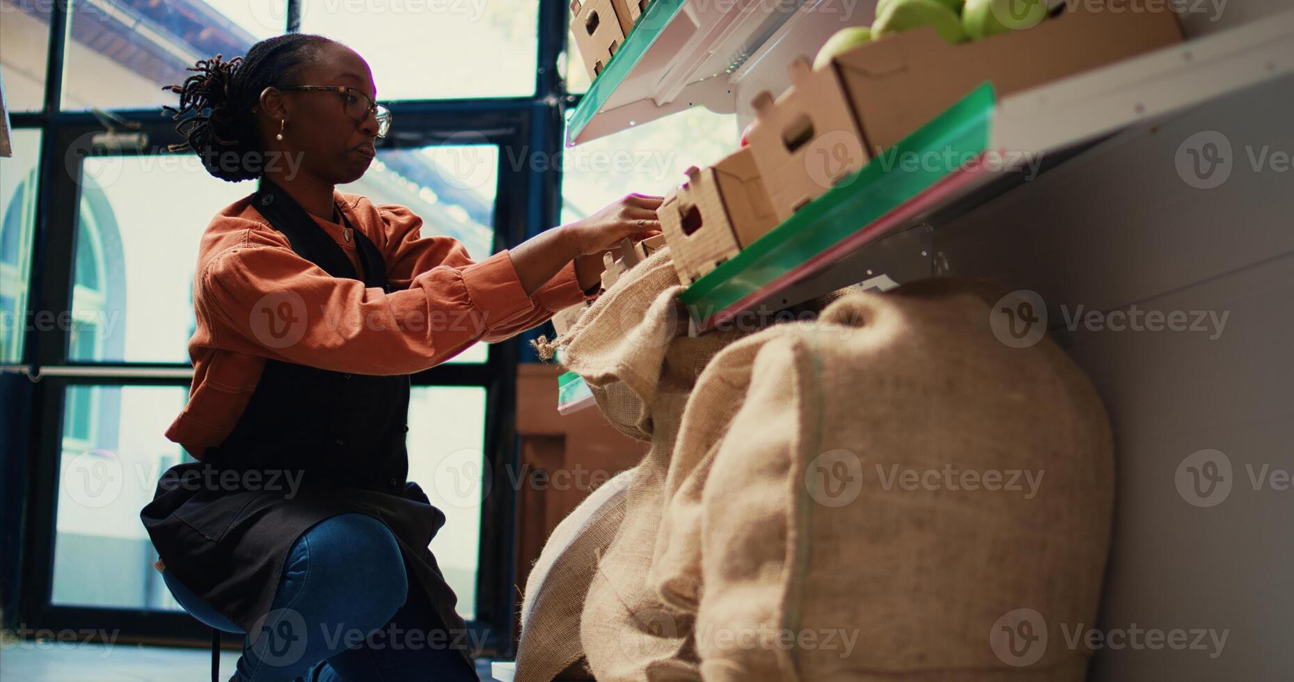 Employee placing crates of organic produce on shelves, preparing store merchandise before welcoming clients. African american woman arranging fruits and vegetables in shop. Handheld shot. photo