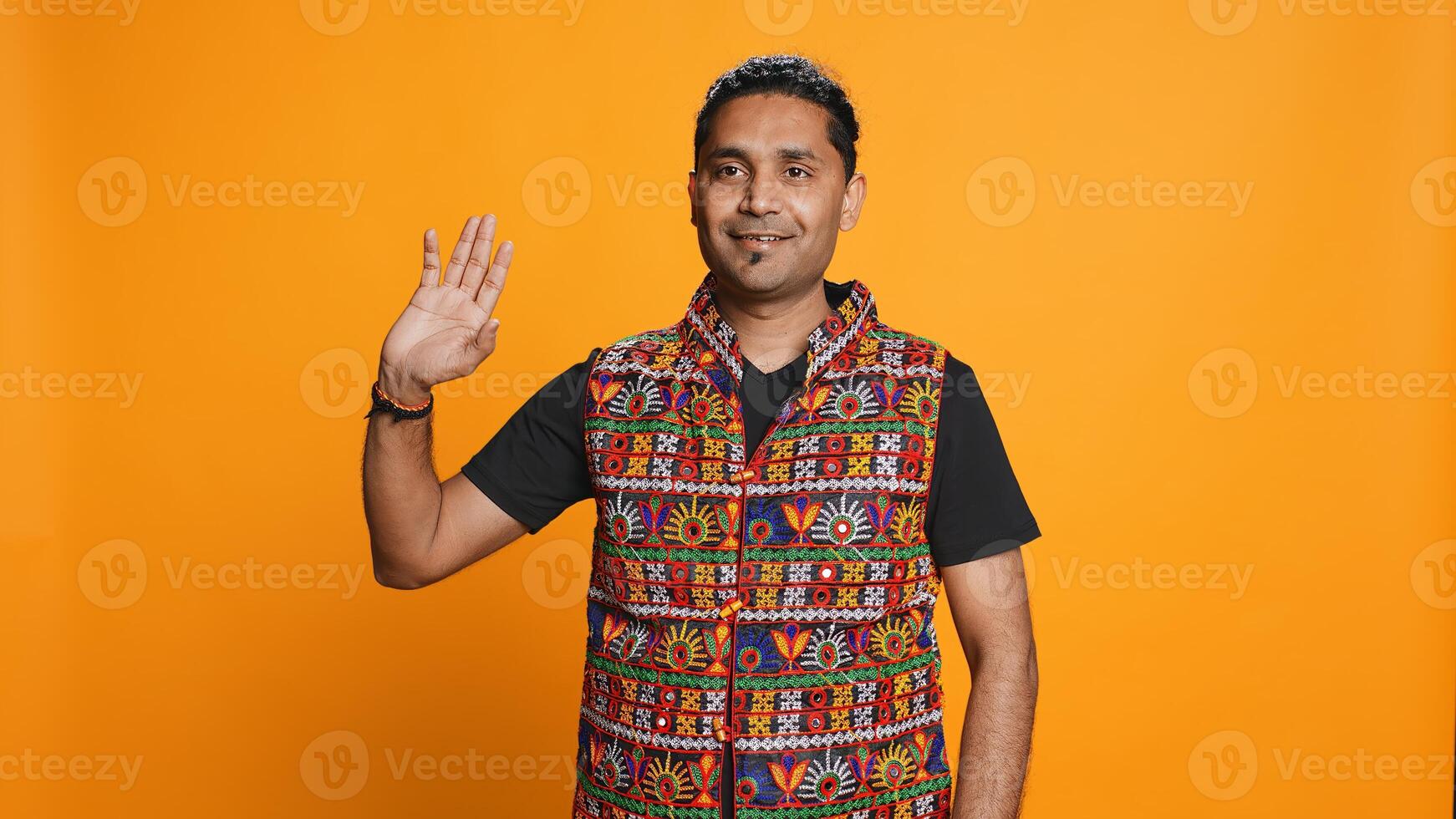 Portrait of jolly man smiling and waving hand, saluting, gesturing, isolated over studio backdrop. Joyous upbeat indian person in traditional attire doing greeting hand gesture, camera B photo