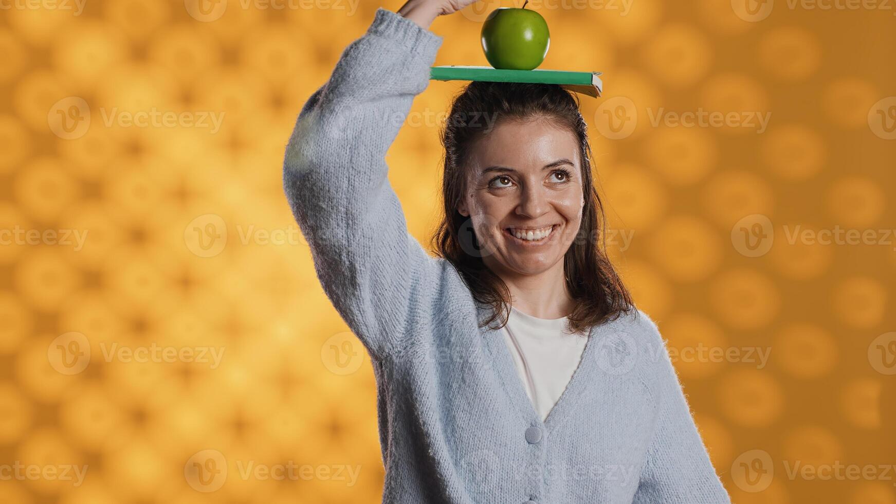 Woman balancing book and apple on head, acting zany, portraying living healthy and having fun, studio background. Bookworm wearing novel and green fruit as hat, enjoying life, goofing around photo