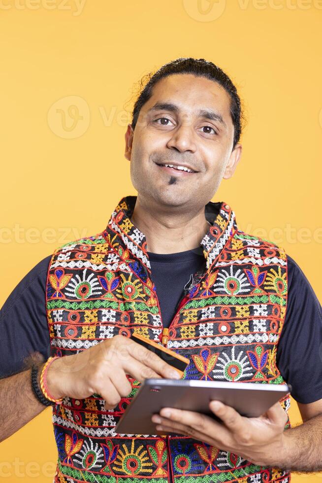 Portrait of jolly man looking at debit card to read banking account, feeling happy to do online purchases, studio background. Cheerful man does internet shopping using digital tablet, having fun photo
