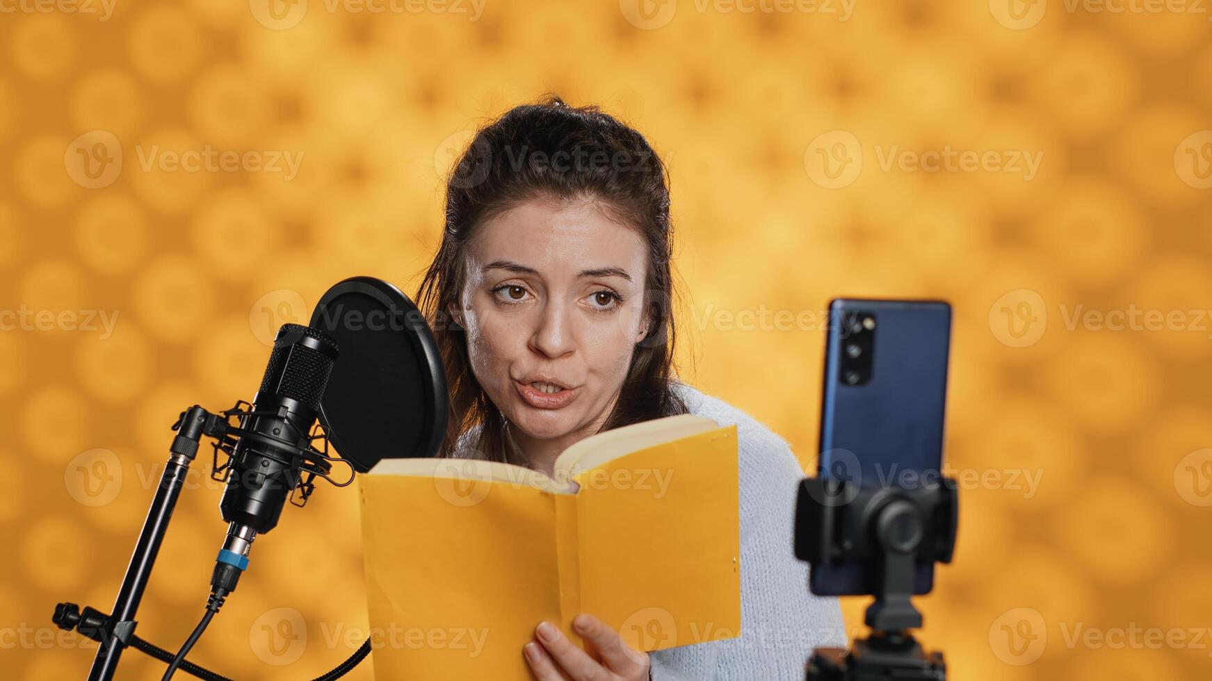 Influencer filming reading book with smartphone on tripod, creating audiobook, studio background. Woman vlogging with mobile phone while producing digital recording on novel using microphone, camera A photo