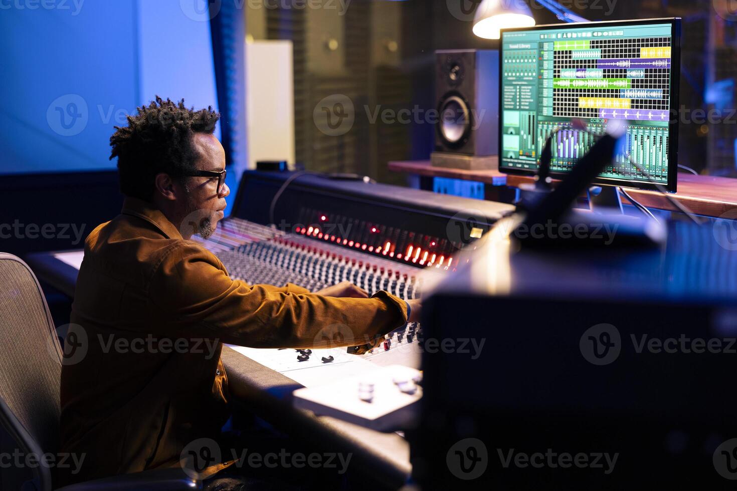 African american record producer composing tunes in control room, editing music and adjusting volume levels with panel board buttons. Audio expert pressing sliders to add sound effects. photo
