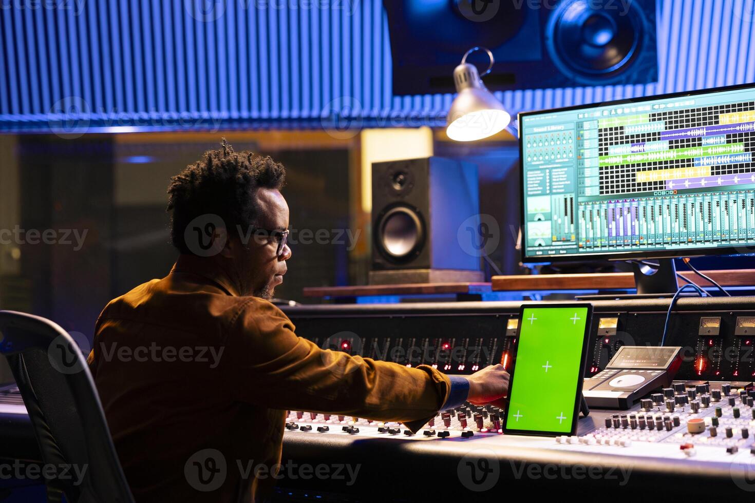 Sound technician mixing tracks next to greenscreen tablet in control room, operating panel board knobs and faders. African american producer recording hit song and processing sounds. photo