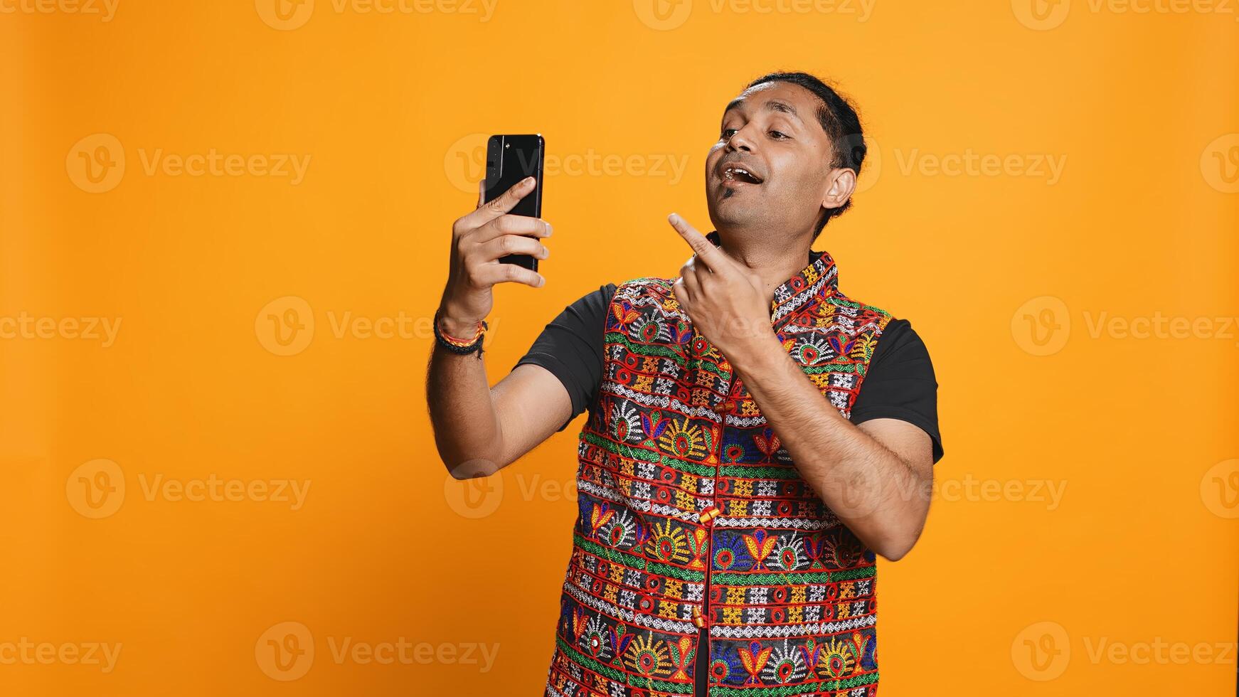 Joyous Indian man greeting friend over online videocall on cellphone, doing friendly gestures. Excited person bonding with mate using mobile phone videoconference, studio background, camera B photo