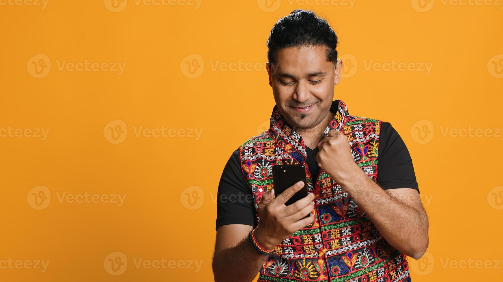 Happy man playing videogames on smartphone, celebrating after defeating foes, doing clenching hand gesture. Player swiping phone screen, thrilled about winning game, studio background, camera A photo