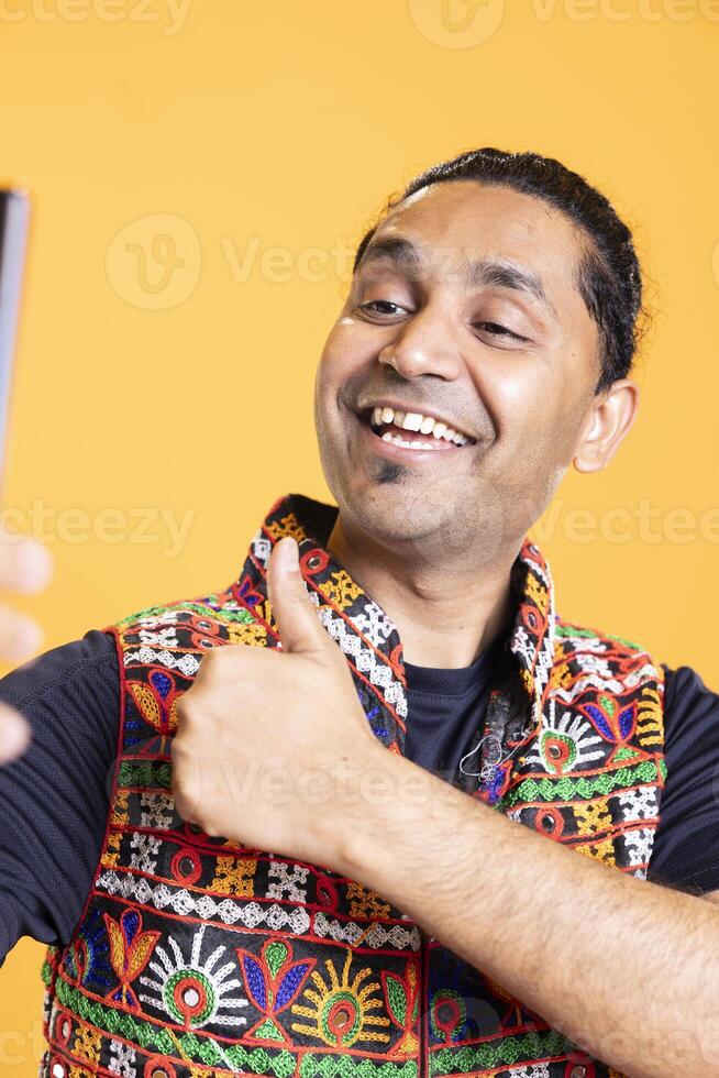 Happy man using cellphone to take selfies, doing thumbs up gesture. Jolly social media user taking photos using phone selfie camera, doing like hand gesturing, studio background