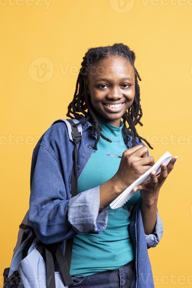 Portrait of happy girl smiling reading and writing for school exam on notepad, studio background. Jolly student grinning, completing papers for research needed for university classes photo