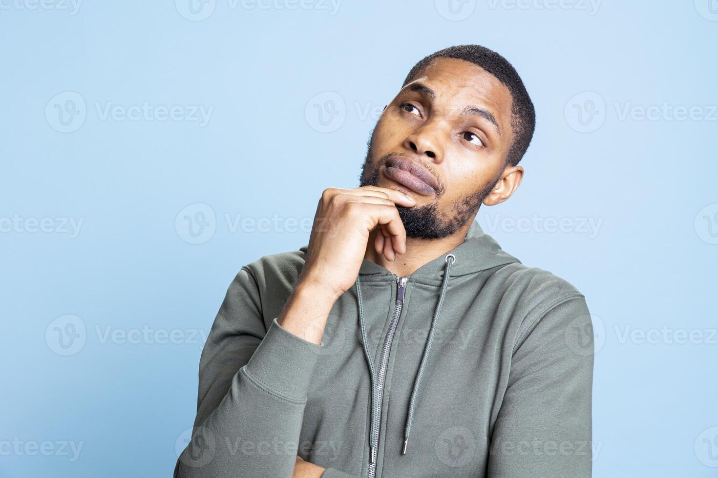 Thoughtful unsure guy with a grey hoodie contemplating about new ideas and solutions for development, being in his thoughts against blue background. African american adult being pensive. photo