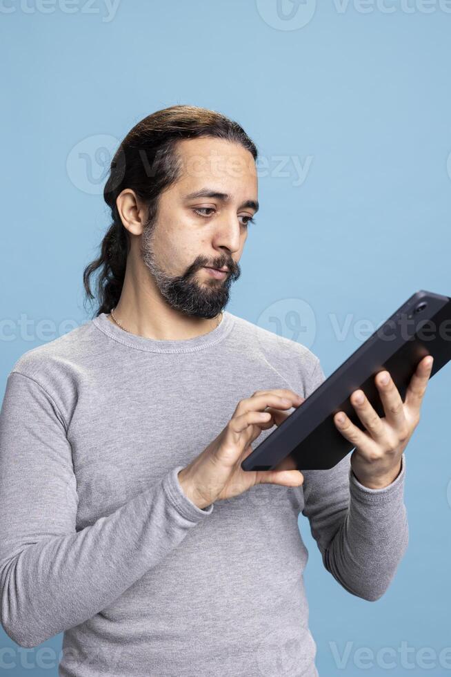 Confident thoughtful man working on his tablet while engaging in digital communication, standing against blue background. Young casual adult doing remote work on modern online platforms. photo