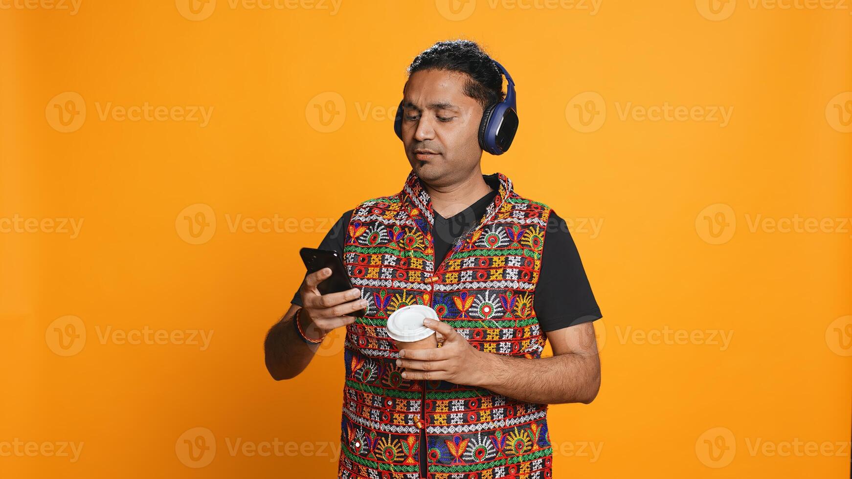 Joyous man listening music, having fun dancing, enjoying hot beverage. Upbeat person wearing headphones, doing funky movements, drinking coffee, studio background, camera B photo