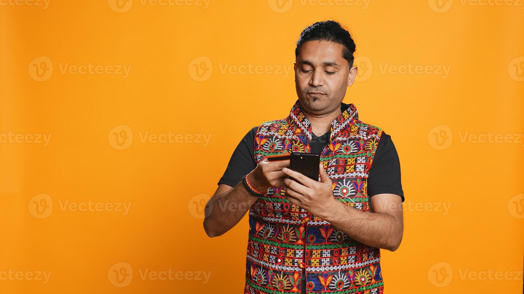 Indian man adding payment method on mobile phone website, purchasing stuff, isolated over studio background. Client does internet shopping, typing debit card info on cellphone, camera B photo
