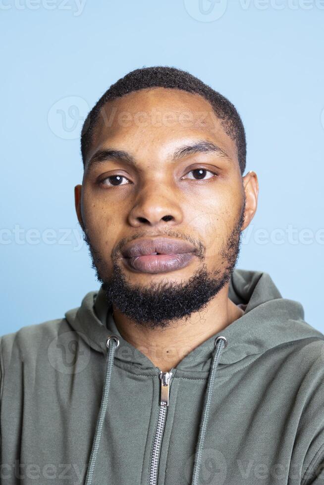 Portrait of african american guy wearing a grey hoodie and smiling, posing in front of the camera and feeling confident. Young positive person being lively and relaxed against blue background. photo