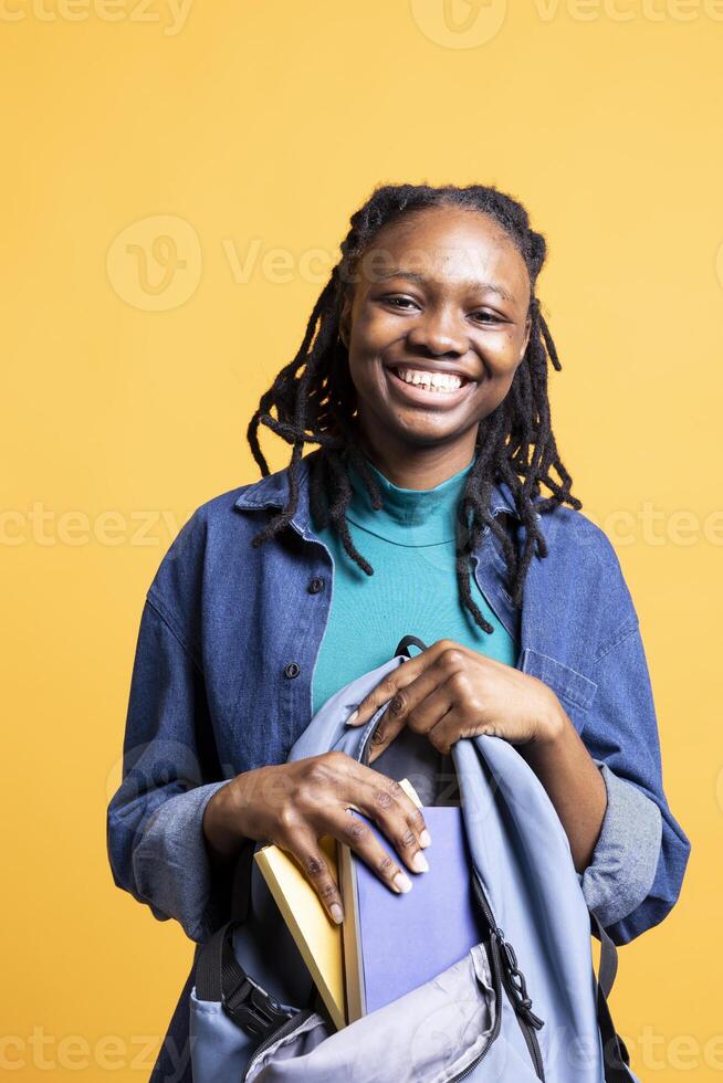 Portrait of happy BIPOC woman browsing through backpack, isolated over studio background. Joyous teenager taking out school textbooks out of rucksack used during commuting to university photo