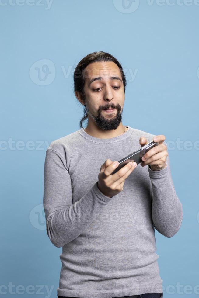 Male gamer having fun with mobile games on his smartphone, enjoying a web tournament on cybersport against blue background. Young adult playing cyber gaming in front of camera. photo