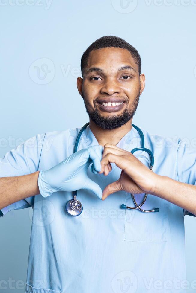 Portrait of cheerful healthcare professional doing heart shape symbol with rubber gloves on hands. Licensed nurse showing love gesture while on duty, standing against blue background. photo