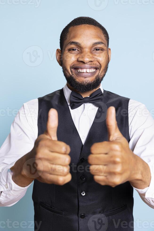 Smiling bellboy in suit and tie showing thumbs up gesture in studio, offering his approval and being satisfied. Positive doorkeeper gives a like sign, trust and professionalism. photo