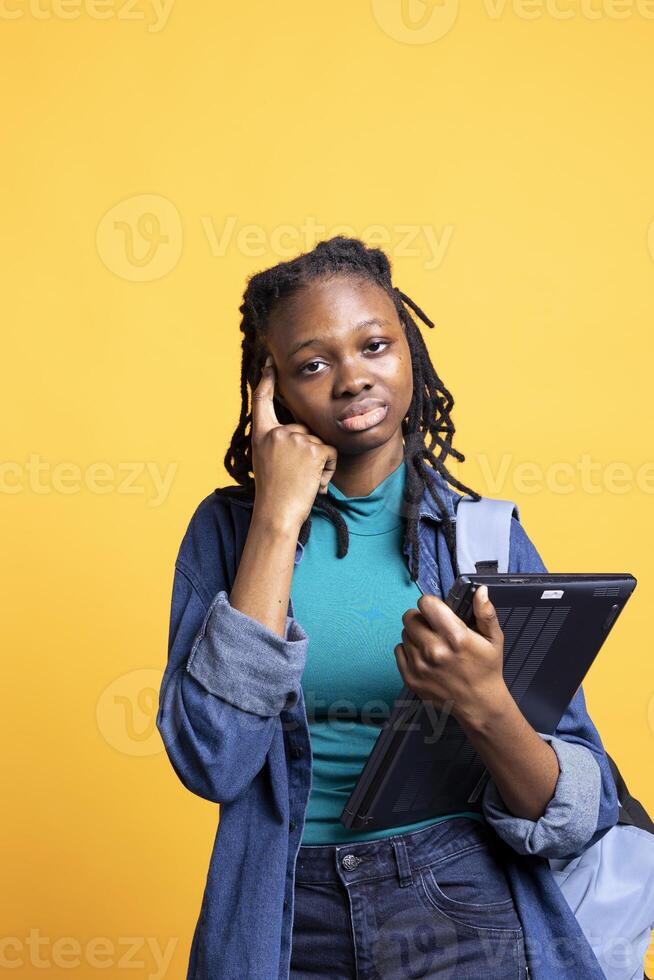 Portrait of BIPOC pensive woman carrying laptop, coming up with idea for school homework, studio background. African american student with notebook doing finger to forehead thinking hand gesture photo