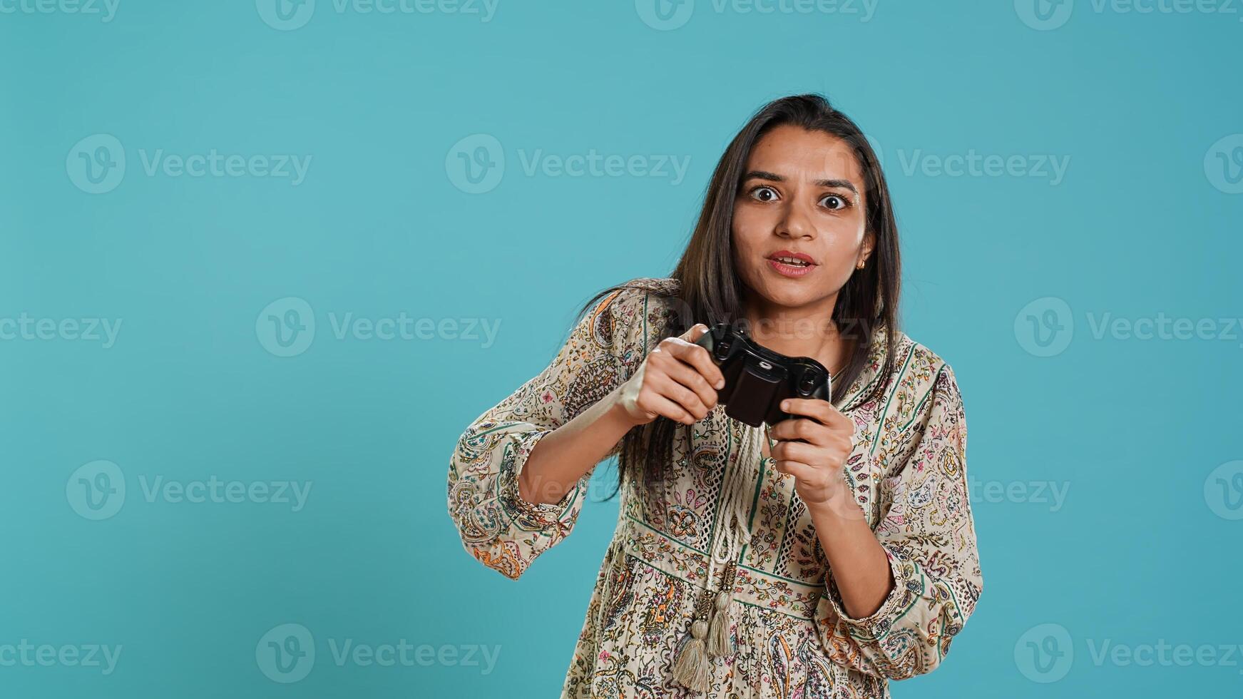 Jolly gamer celebrating after winning game on gaming console, studio background. Delighted Indian woman bragging after being victorious in videogame, defeating all enemies using gamepad, camera A photo