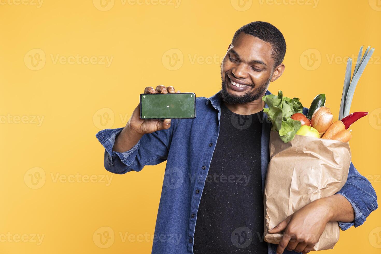 Happy charismatic man presenting greenscreen layout on camera, carrying a bag of groceries and smiling. Confident vegan person showing isolated mockup template on phone display. photo