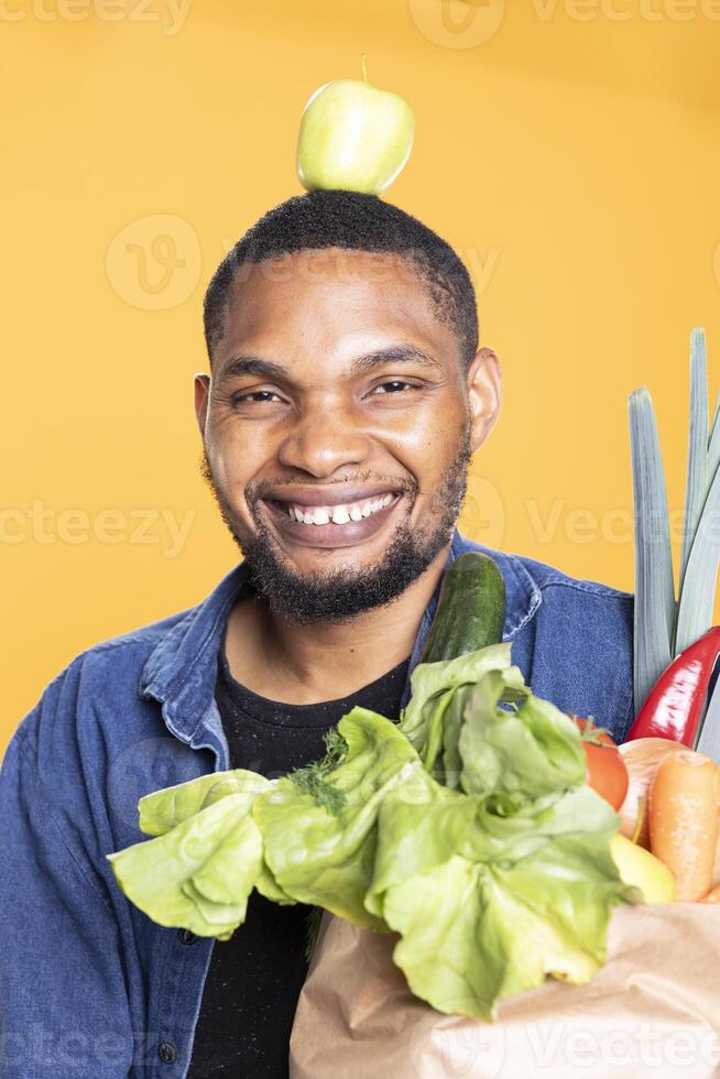African american man poses with a green apple on his head in studio, having fun with fresh ethically sourced fruits. Charismatic vegan guy holding a bag of eco friendly groceries, zero waste. photo