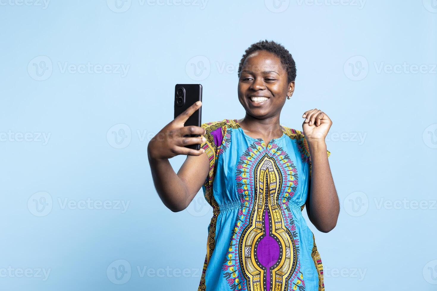 African american person having fun with photographs on smartphone, smiling at the camera and taking pictures in studio. Optimistic positive woman enjoy taking photos in ethnic attire.
