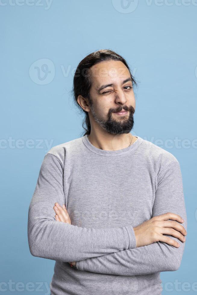 Sweet playful person winking and making jokes against blue background, acting casual with a vibrant confident attitude. Young cheerful guy being flirty and cool, self assured man. photo