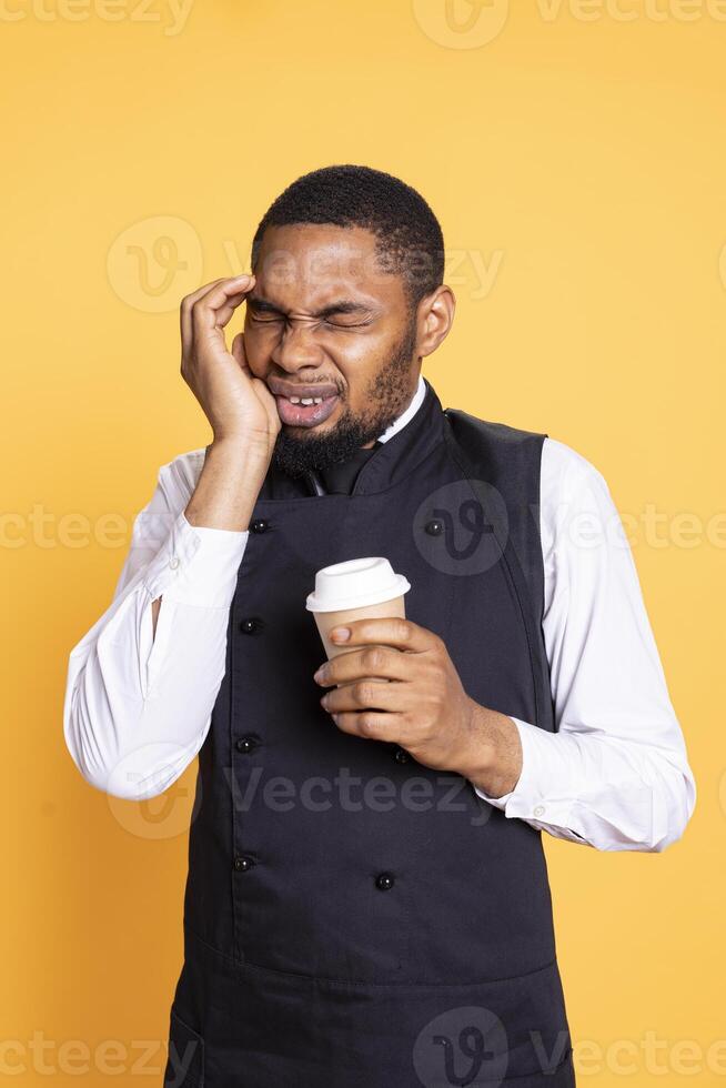 Restaurant worker suffering from a painful migraine while he serves people, drinking a cup of coffee to stay awake. Unwell butler feeling sick with a headache, burnout and fatigue. photo