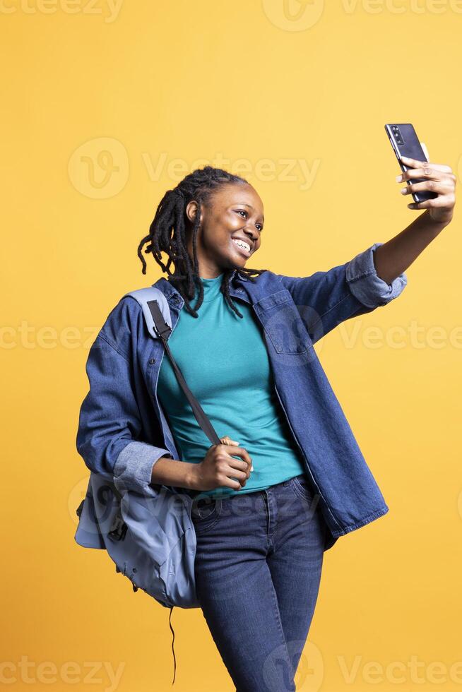Cheerful woman using smartphone to take selfies and post them on social media. Happy BIPOC person taking photos using phone selfie camera, isolated over yellow studio background