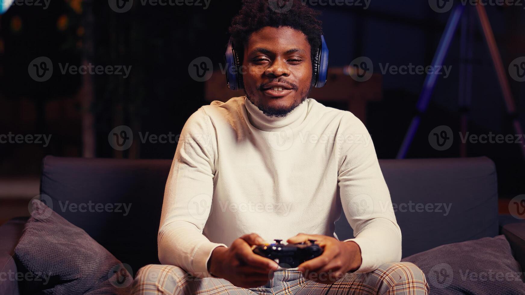 African american man having online gaming session with friends, talking with them through headphones microphone. Gamer playing videogames with teammates on console using gamepad controller photo