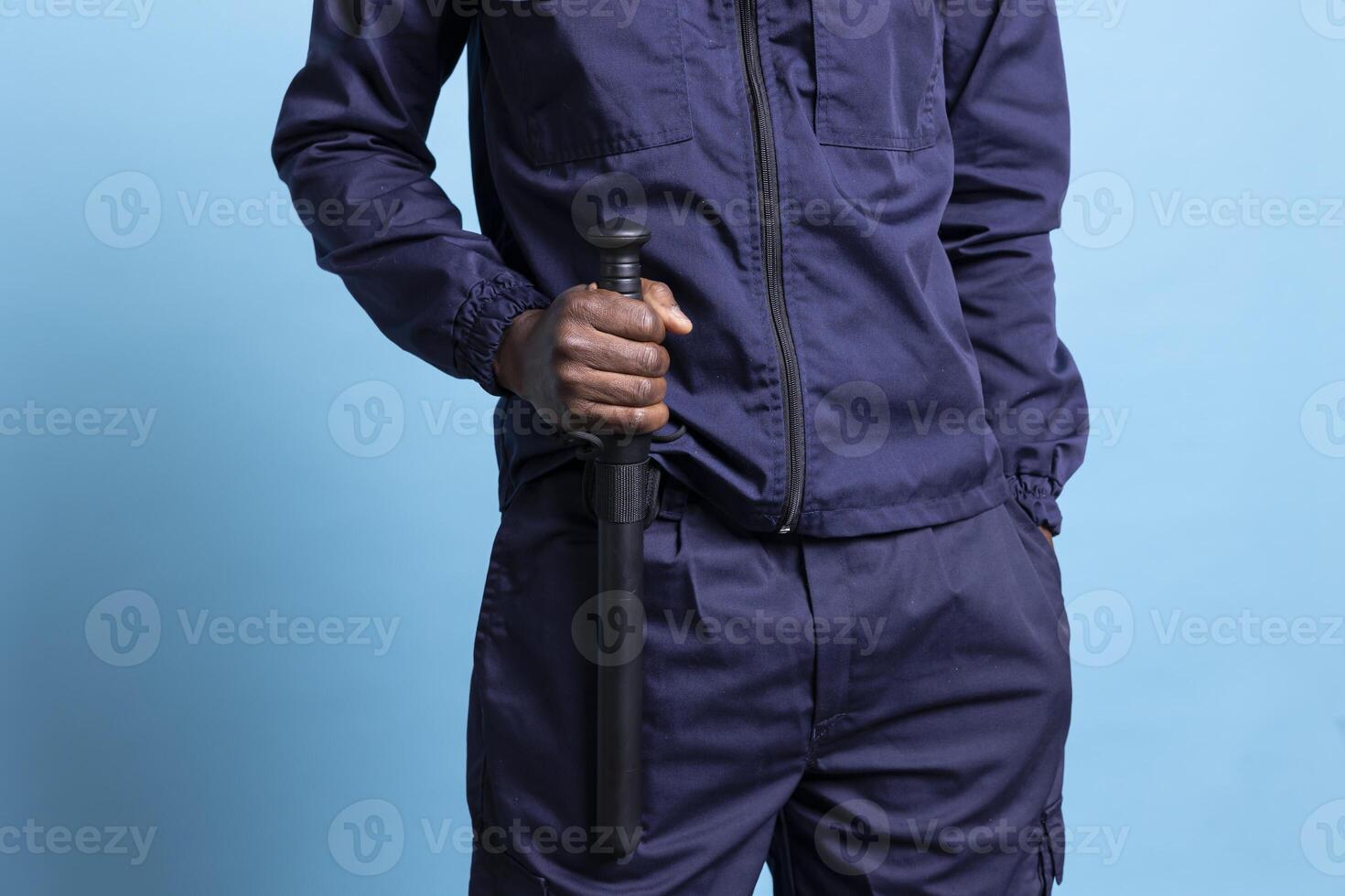 Security agent holding his baton for own safety while on duty, safeguarding and protecting the premises. Trained young bodyguard in work uniform staying alert against blue background. Close up. photo