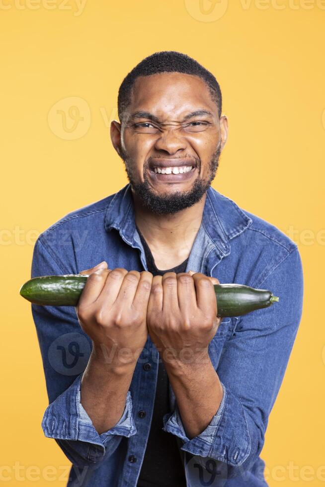 African american male model playing around with organic veggies, to lifting weight with a ripe cucumber against yellow background. Vegan person fooling around with fresh produce. photo
