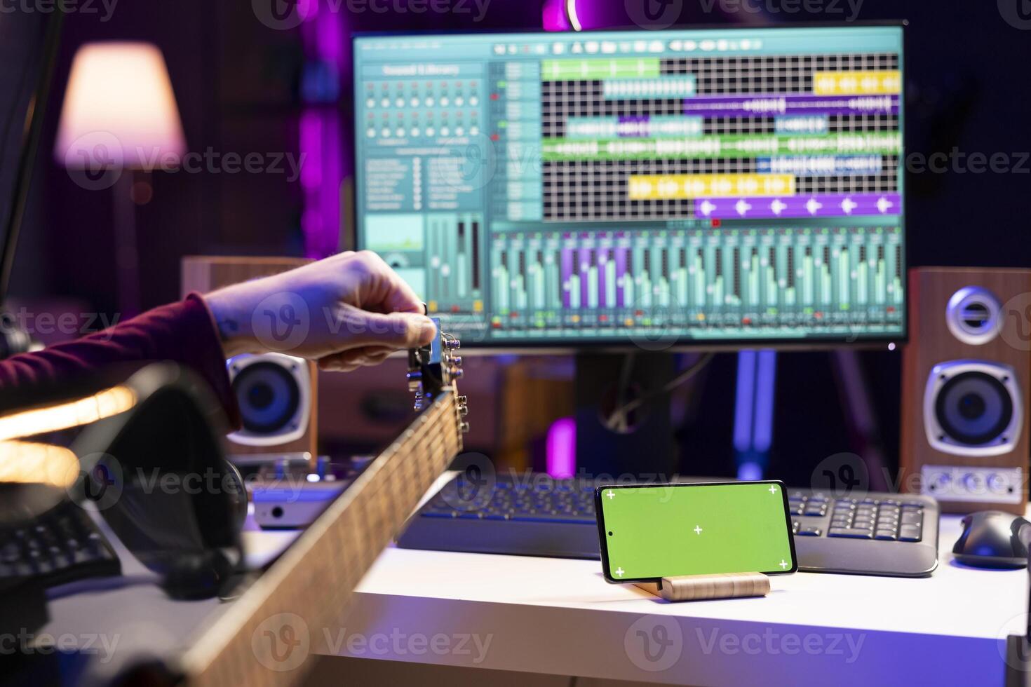 Beginner guitarist watching online tutorial to learn tuning his guitar strings, looks at greenscreen display on mobile phone. Artist learning to play acoustic instrument, practicing new accords. photo