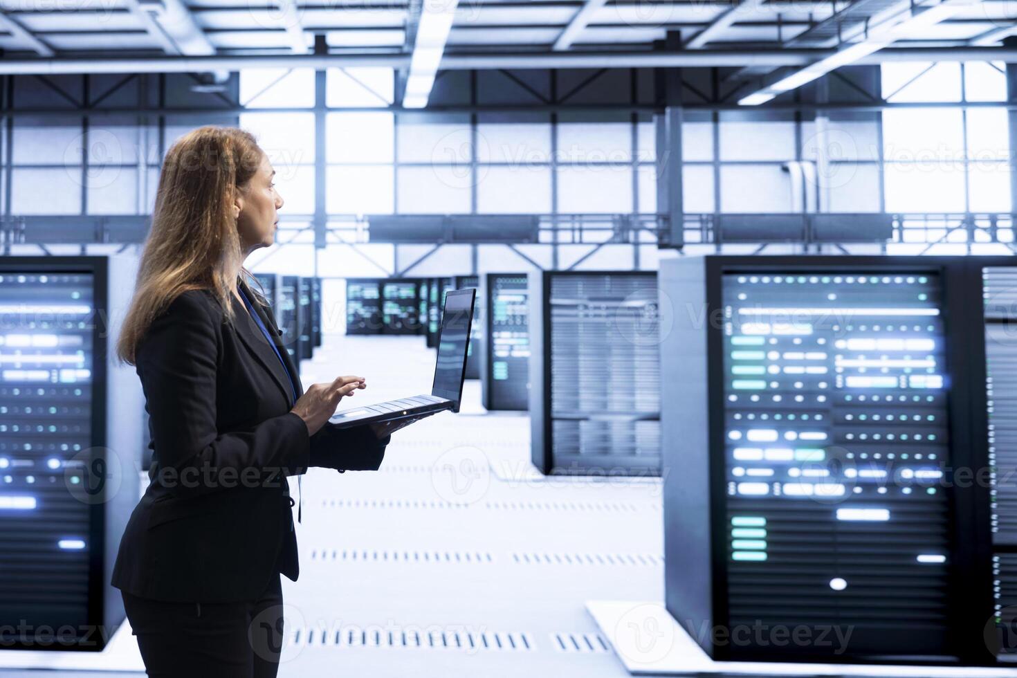Computer scientist in data center checking rigs and hardware, doing checkup. Woman walking in high tech facility, planning and executing server hub migrations and upgrades using notebook photo