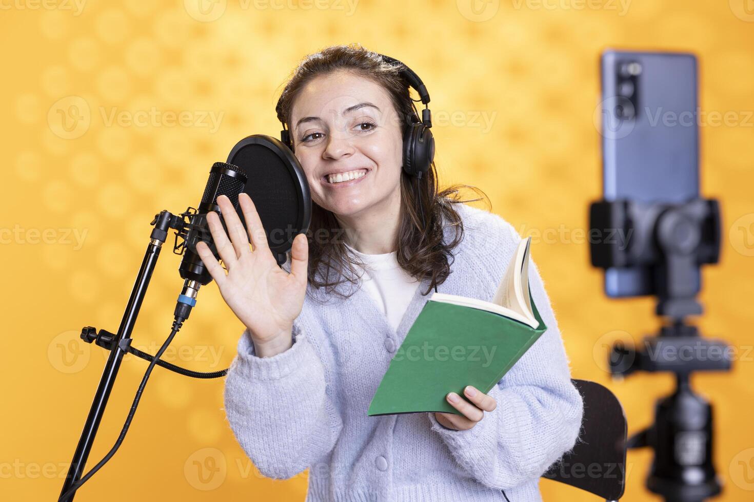 Happy media star waving hand while filming vlog at work, reading book to record audiobook. Cheerful woman doing content creation, recording novel, studio background, doing greeting gesture photo