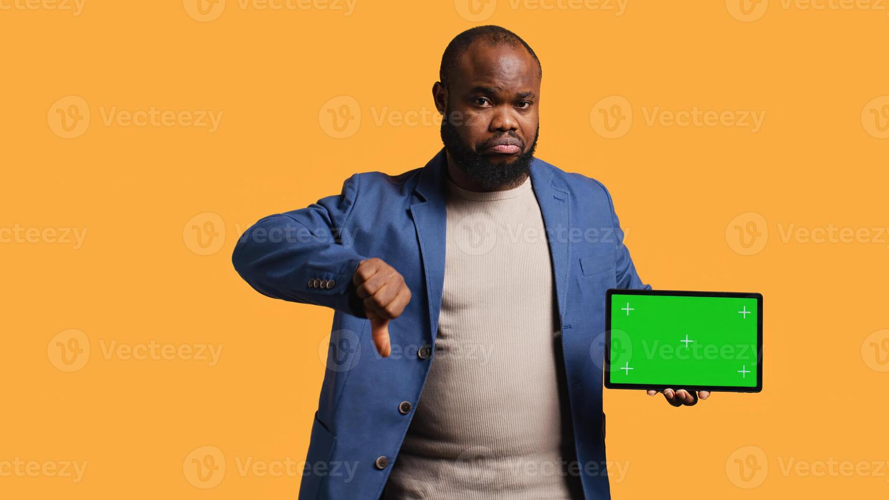 Upset african american man shows thumbs down sign gesturing holding isolated screen tablet, disapproving. BIPOC person does rejection hand gesture regarding mockup device, studio background, camera B photo