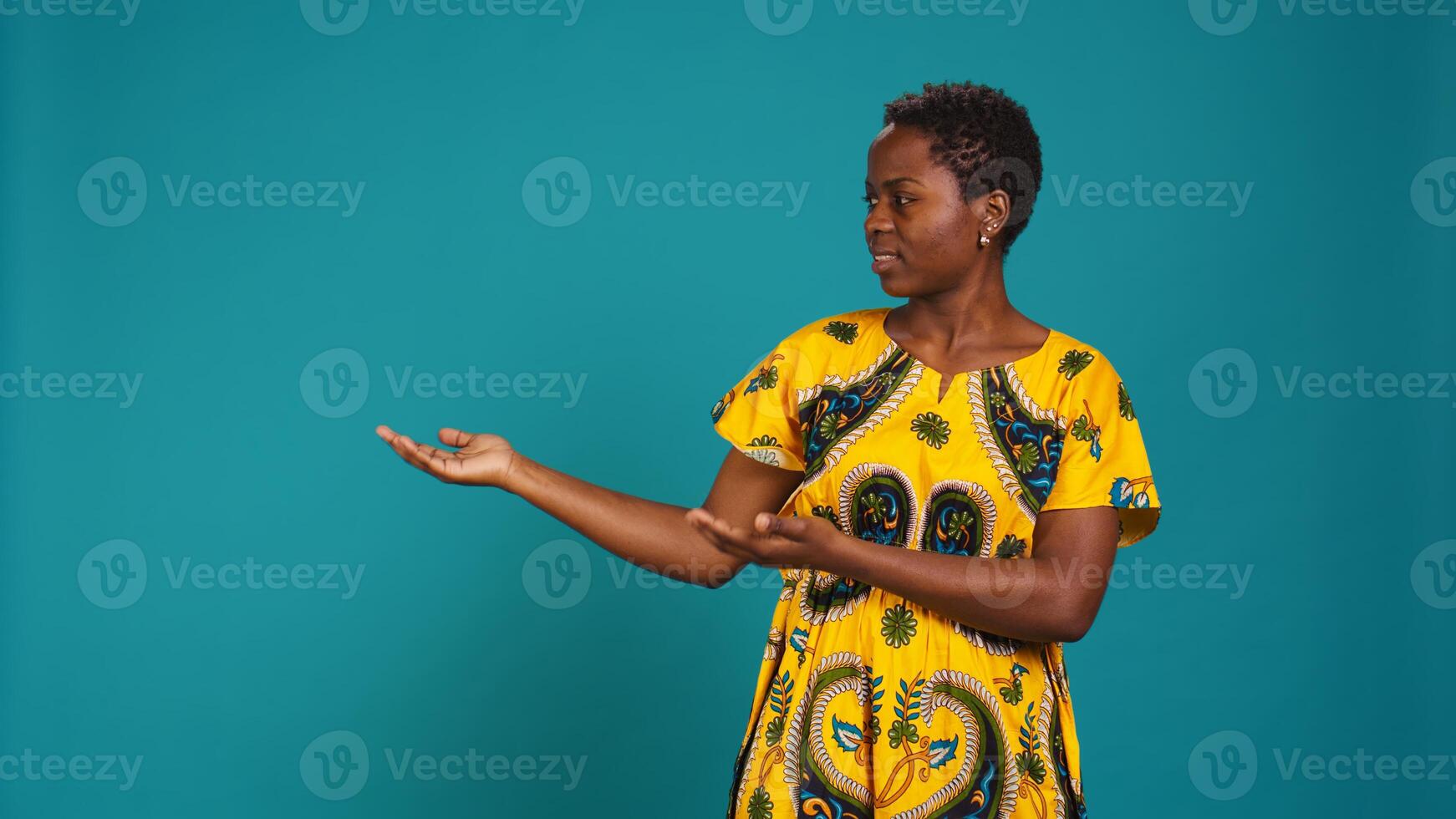 Confident smiling girl indicating direction to left or right sides in studio, pointing her index fingers around. Young natural person wears traditional african attire with tribal native elements. Camera B. photo
