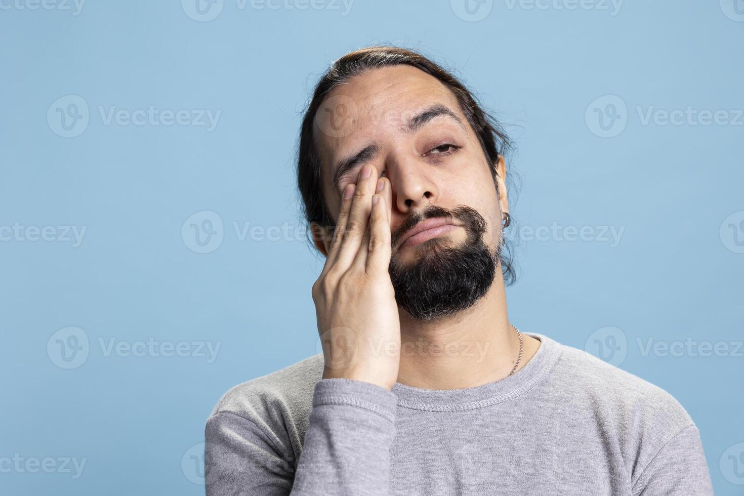 Middle eastern exhausted overwhelmed guy deals with fatigue in studio, being extremely tired and almost falling asleep against blue background. Young adult feels sleepy and overworked. photo
