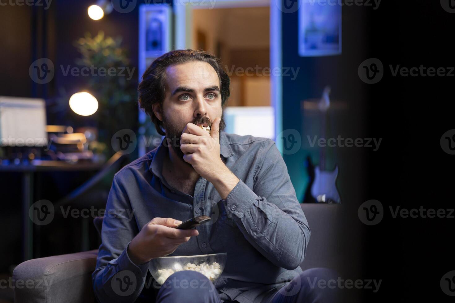 Cord cutter at home looking in disbelief at TV displaying thriller film, stuffing mouth with popcorn. Stupefied person at home watching suspenseful movie on VOD channel, perplexed by plot twist photo