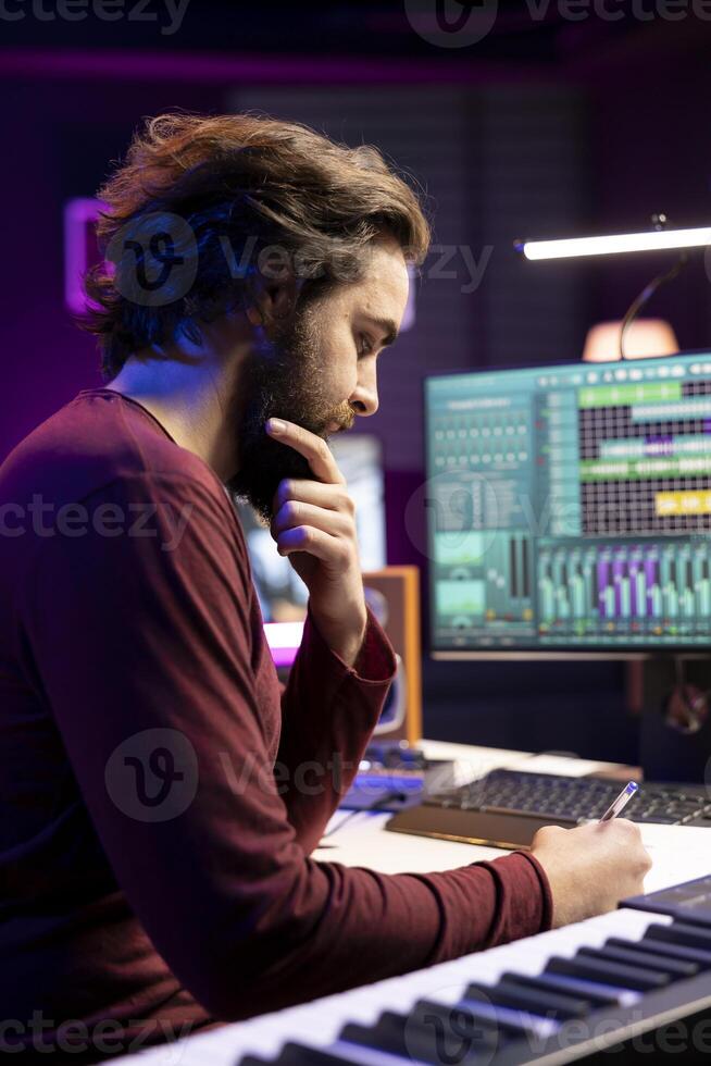 Professional songwriter writes down new verses and rhymes in home studio, creating new music with daw software. Young artist works with audio equipment and gear, recording tunes. photo