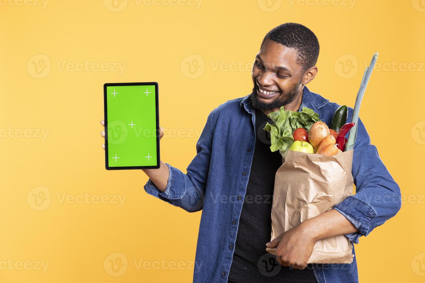 Confident african american man presents greenscreen on a tablet, carrying bag of vegetables against yellow background. Pleased male model showing isolated mockup display on device. photo