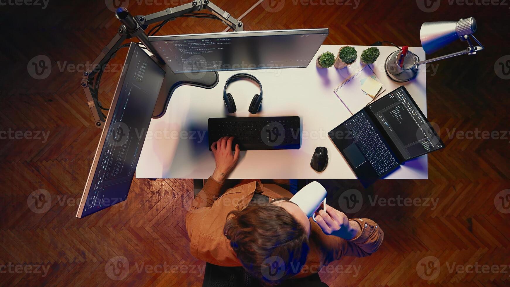 Top down view of engineer doing multitasking to finish developing PC code, drinking coffee, listening music. IT admin wearing headphones, solving database errors on computer and laptop, aerial shot photo