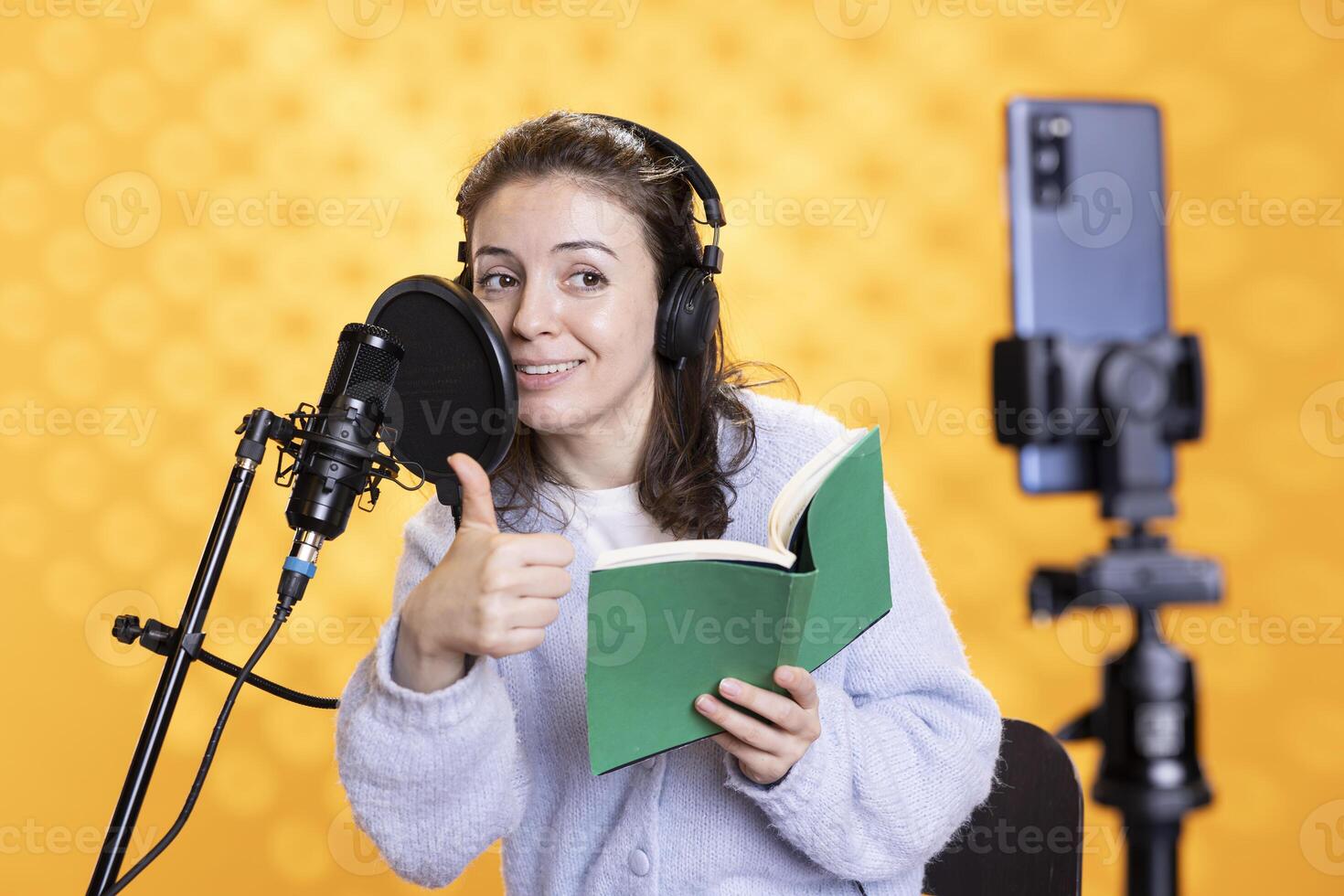 Smiling influencer doing thumbs up at phone camera, filming reading book, creating audiobook, studio background. Influencer showing positive hand gesture, doing vlog while recording audio novel photo