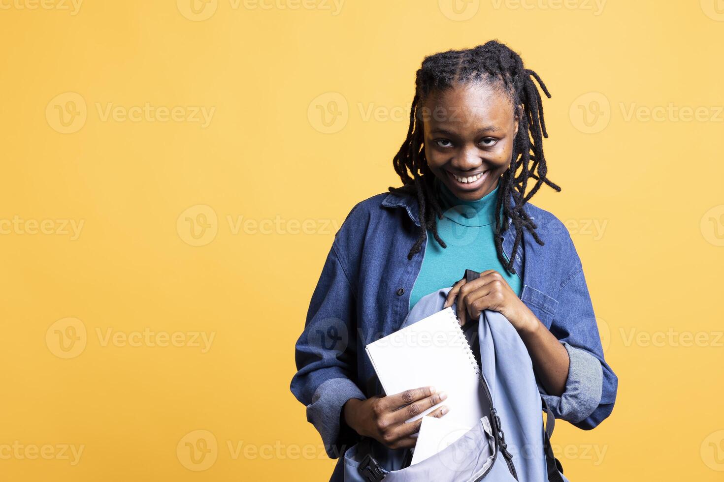 Portrait of smiling woman rummaging through backpack, isolated over yellow studio background. Cheerful girl taking out school supplies out of rucksack used during commuting to university photo