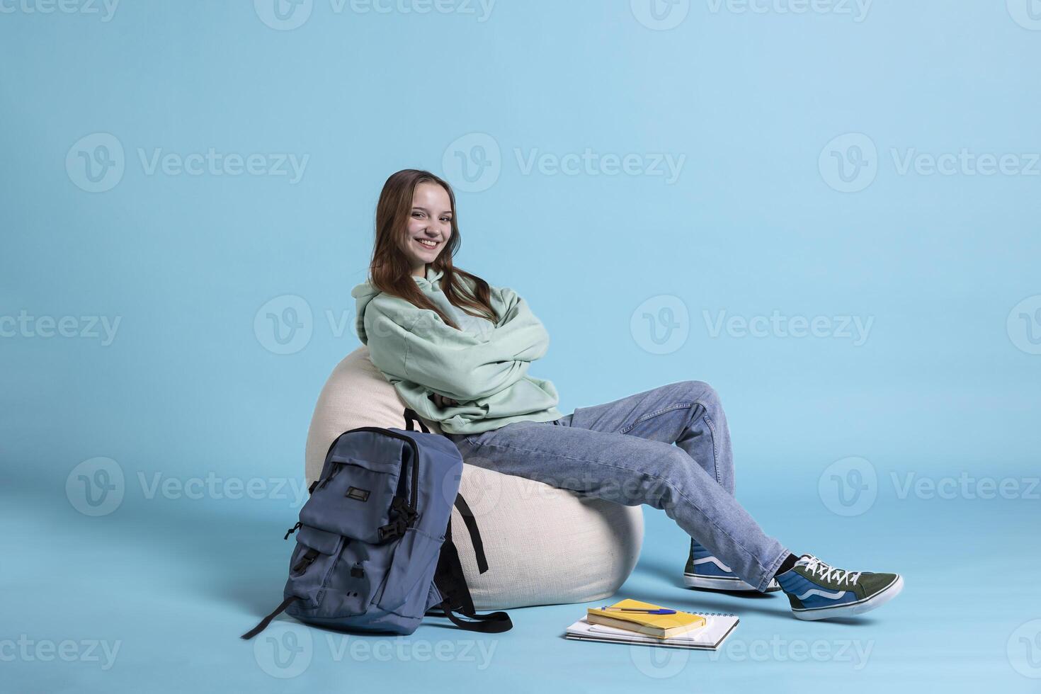 Portrait of radiant teenager with rucksack grinning, holding school notebook and notes, studio backdrop. Upbeat schoolgirl with textbook in arms, preparing to go to high school photo