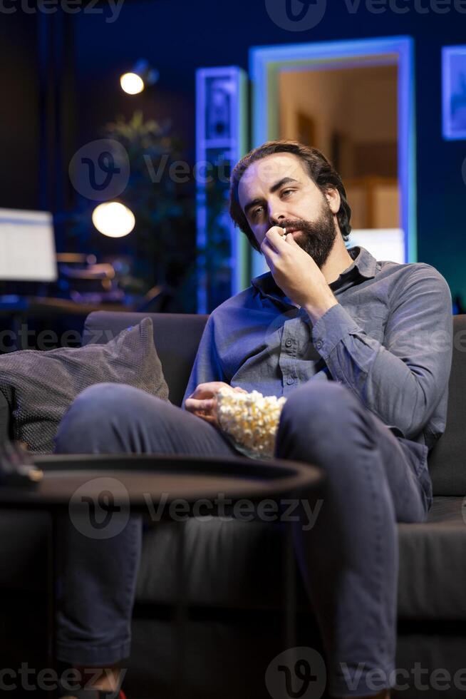 Fatigued man watching shows on TV and munching on popcorn, paying close attention to screen. Tired sedentary man sitting in pajamas all day devouring snacks and consuming mindless entertainment photo