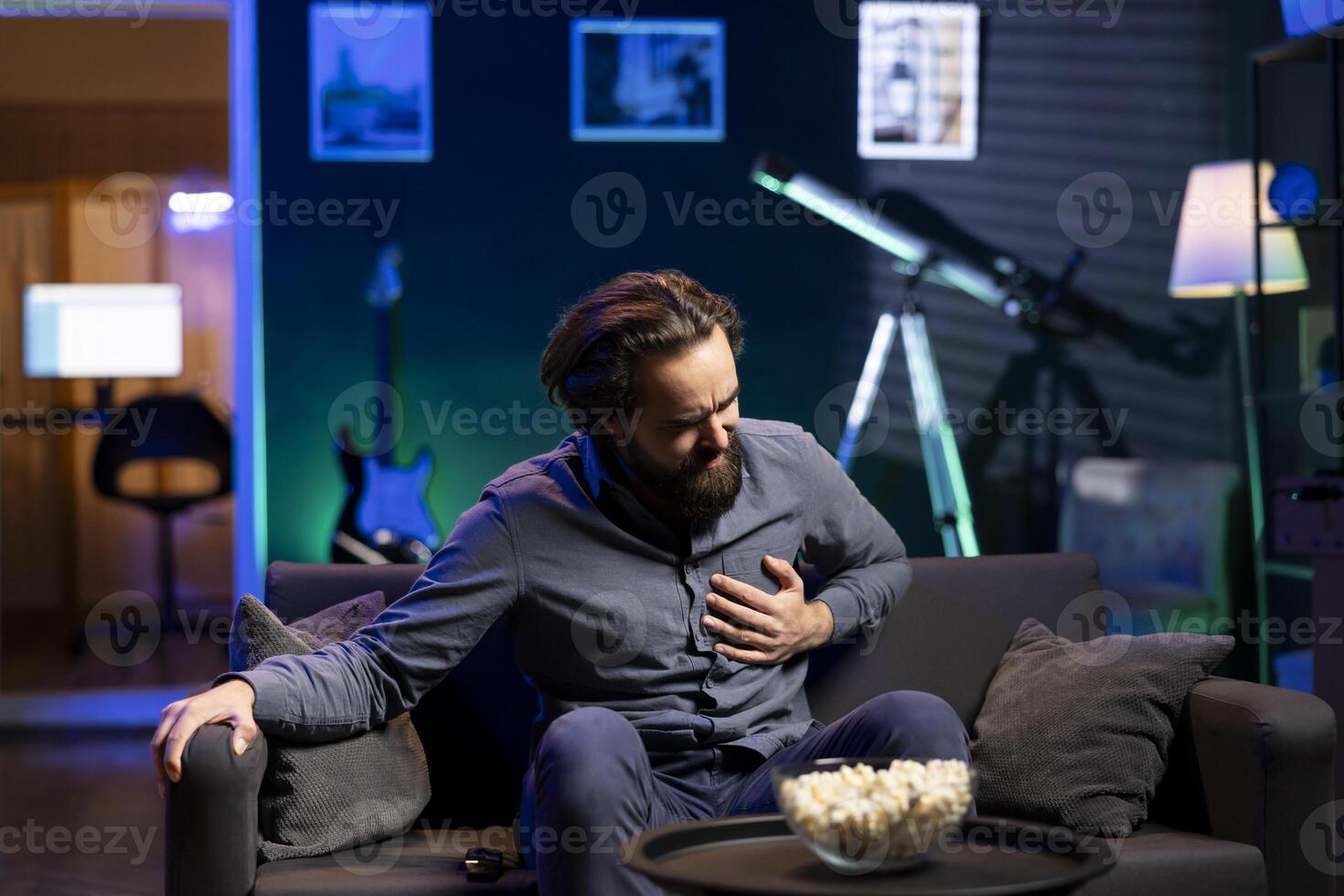 Sedentary man at home feeling terrible pain in chest while watching TV on couch after lack of activity. Person getting painful cardiac stroke from consuming unhealthy snacks all day, gripping chest photo