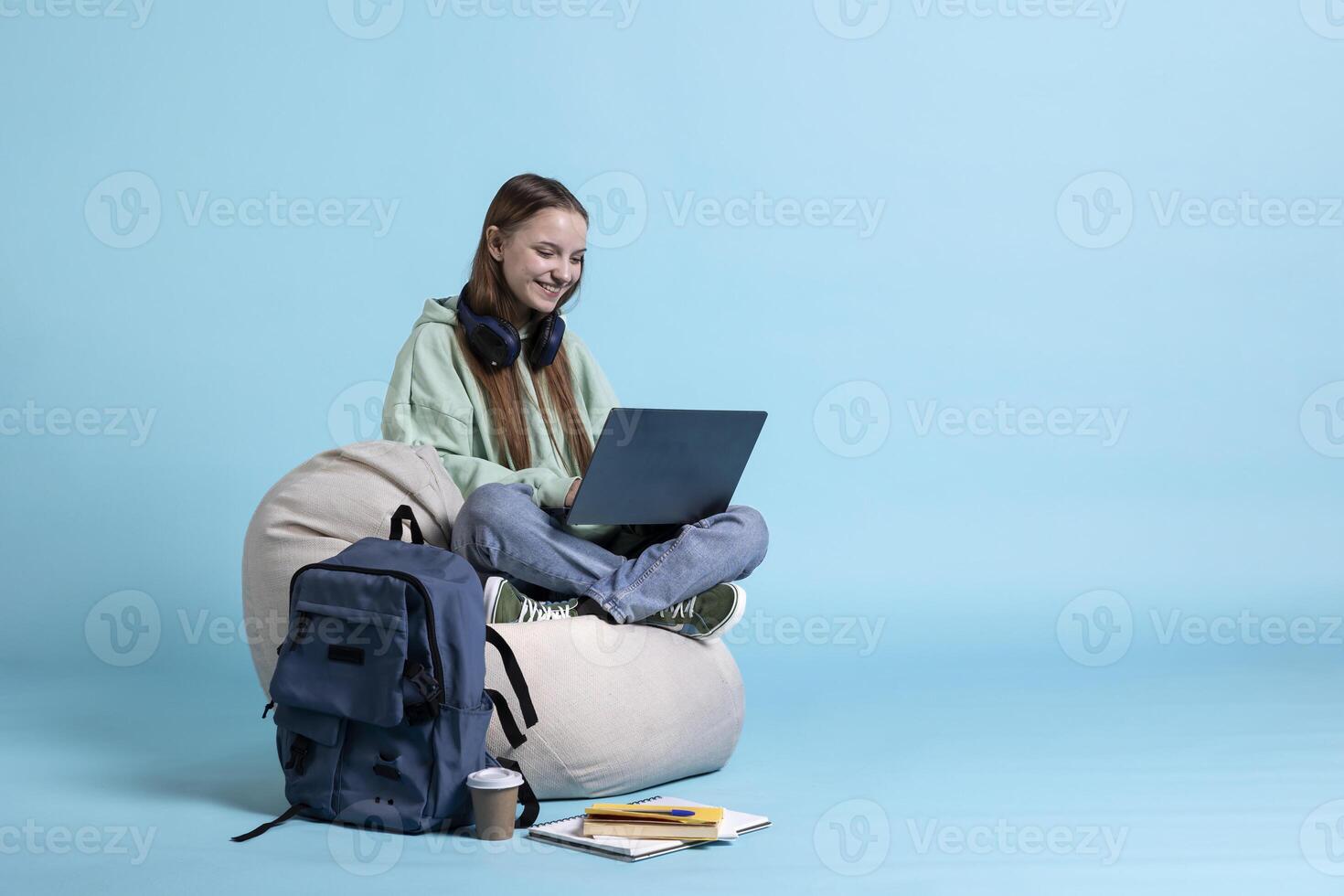 Smiling woman scrolling on laptop to check social media feed, relaxing on beanbag. Happy teenager browsing internet websites on notebook, isolated over blue studio background photo